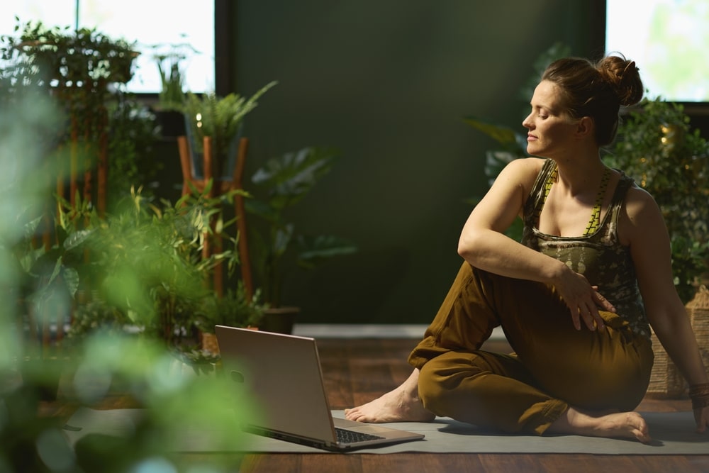 Woman in Yoga pose sits in tranquil room with dark green walls, plants