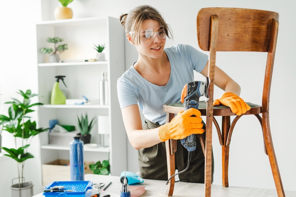 Woman upcycling, refinishing wooden chair with goggles in minimalist sitting room with plants